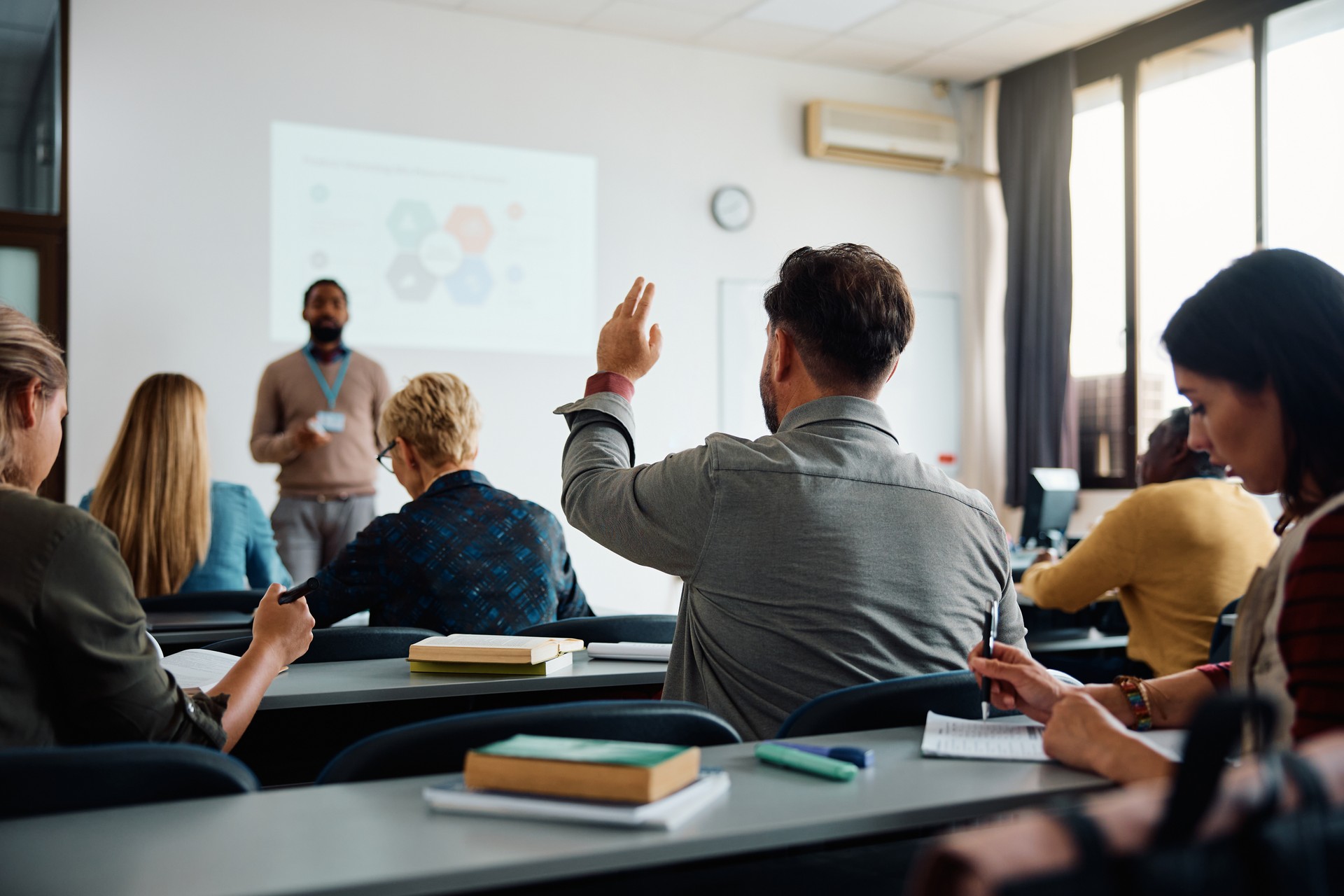 Nexodus Back view of student raising his hand to answer teacher's question during education training class.