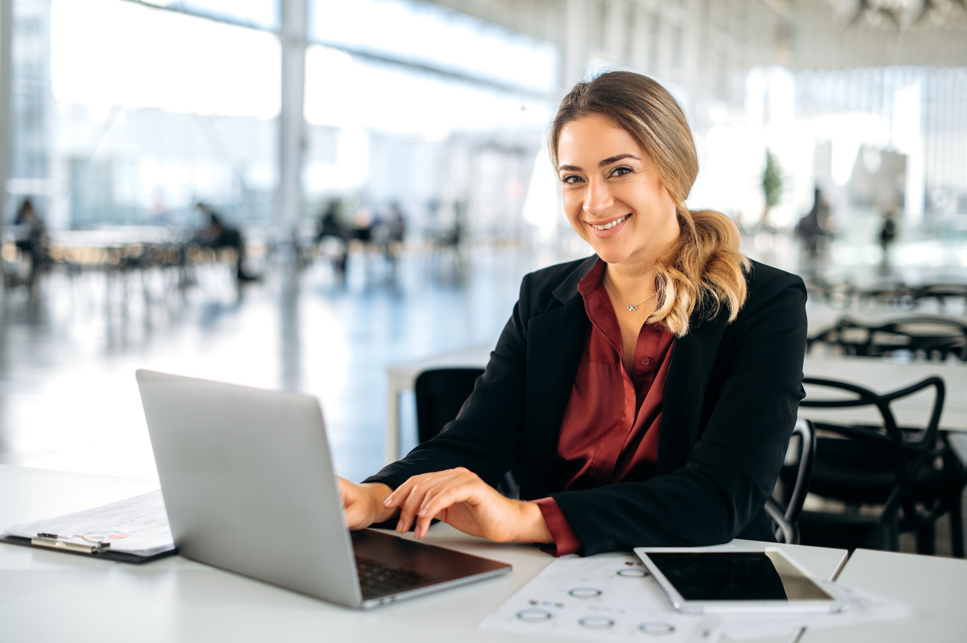 Nexodus Photo of a confident lovely elegantly dressed positive caucasian business lady, executive, recruitment, product manager, sits in a business center, working in a laptop, looks at camera, smile friendly